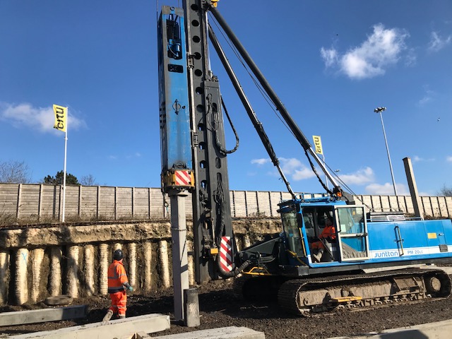 Rotary piling rig driving a pile beside a site boundary wall
